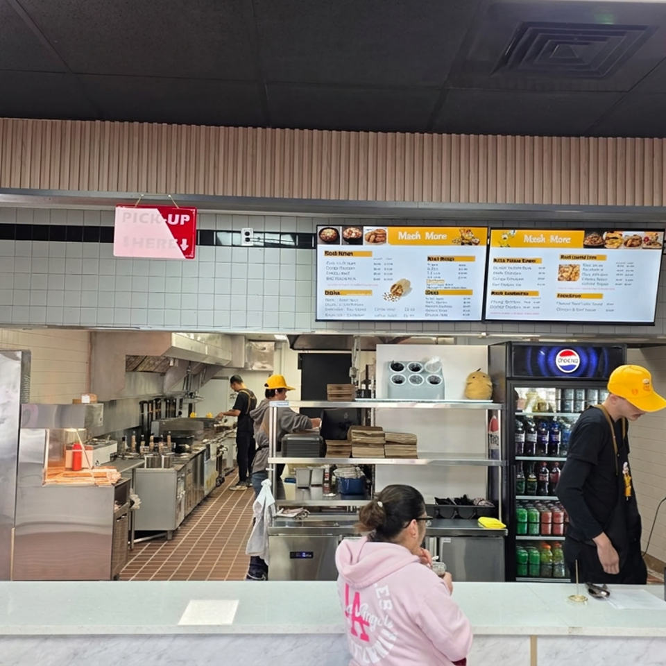 Restaurant counter with staff serving customers beneath digital menus, featuring tiled walls, pickup and order signs, and a beverage cooler.